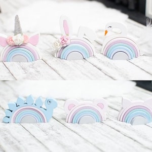 May include: Set of six rainbow wooden stacking toys with different animal toppers. The toppers include a unicorn, bunny, swan, dinosaur, bear, and a cloud. The rainbows are painted in pastel pink, blue, and white.