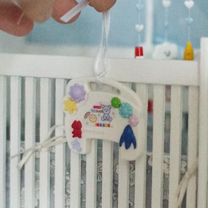 May include: A miniature nursery scene with a white crib, dresser, and shelves. A colorful mobile hangs above the crib. The shelves hold decorative items, including a cactus and a bunny figurine. The room is painted in light blue.