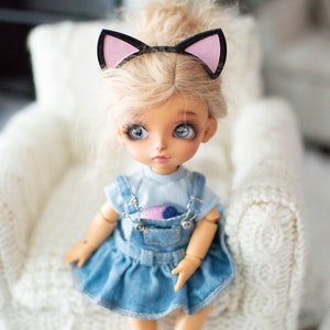 May include: A doll with blonde hair and blue eyes wearing a denim dress and a cat ear headband. The doll is sitting on a white, textured chair. The doll's outfit includes a light blue top and a denim overall dress.