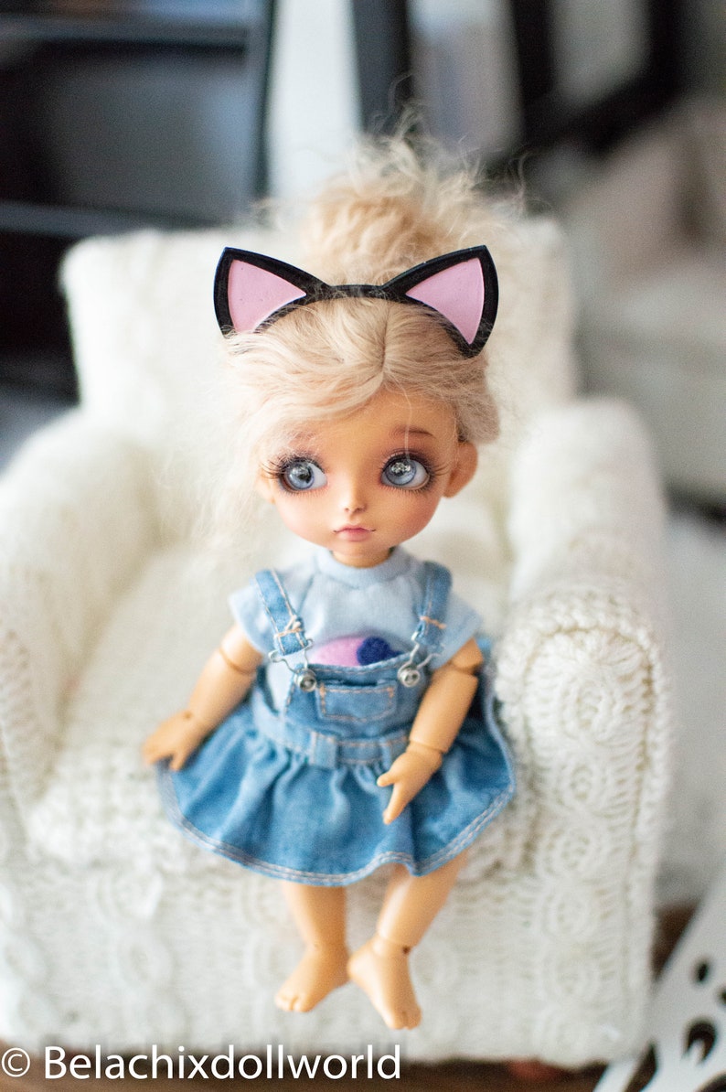 May include: A doll with blonde hair and blue eyes wearing a denim dress and a cat ear headband. The doll is sitting on a white, textured chair. The doll's outfit includes a light blue top and a denim overall dress.