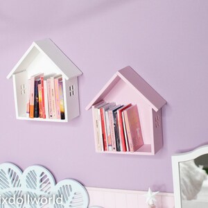 May include: Two white and pink wooden house-shaped shelves with books on them, mounted on a light purple wall.