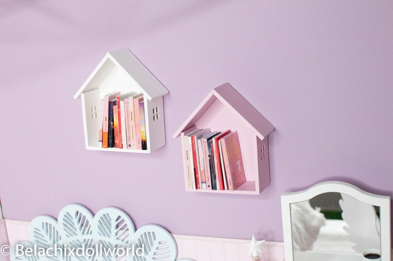 May include: Two white and pink wooden house-shaped shelves with books on them, mounted on a light purple wall.