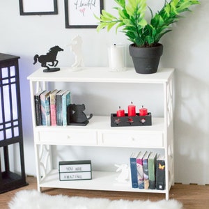 May include: White bookshelf with three shelves, two drawers, and decorative accents. The top shelf has a black horse silhouette, a white horse silhouette, a potted green plant, and a white candle. The middle shelf has a black cat bookend, a set of red candles in a black holder, and a stack of books. The bottom shelf has a black light-up sign that says "You Are Amazing", a white rabbit figurine, and a stack of books.