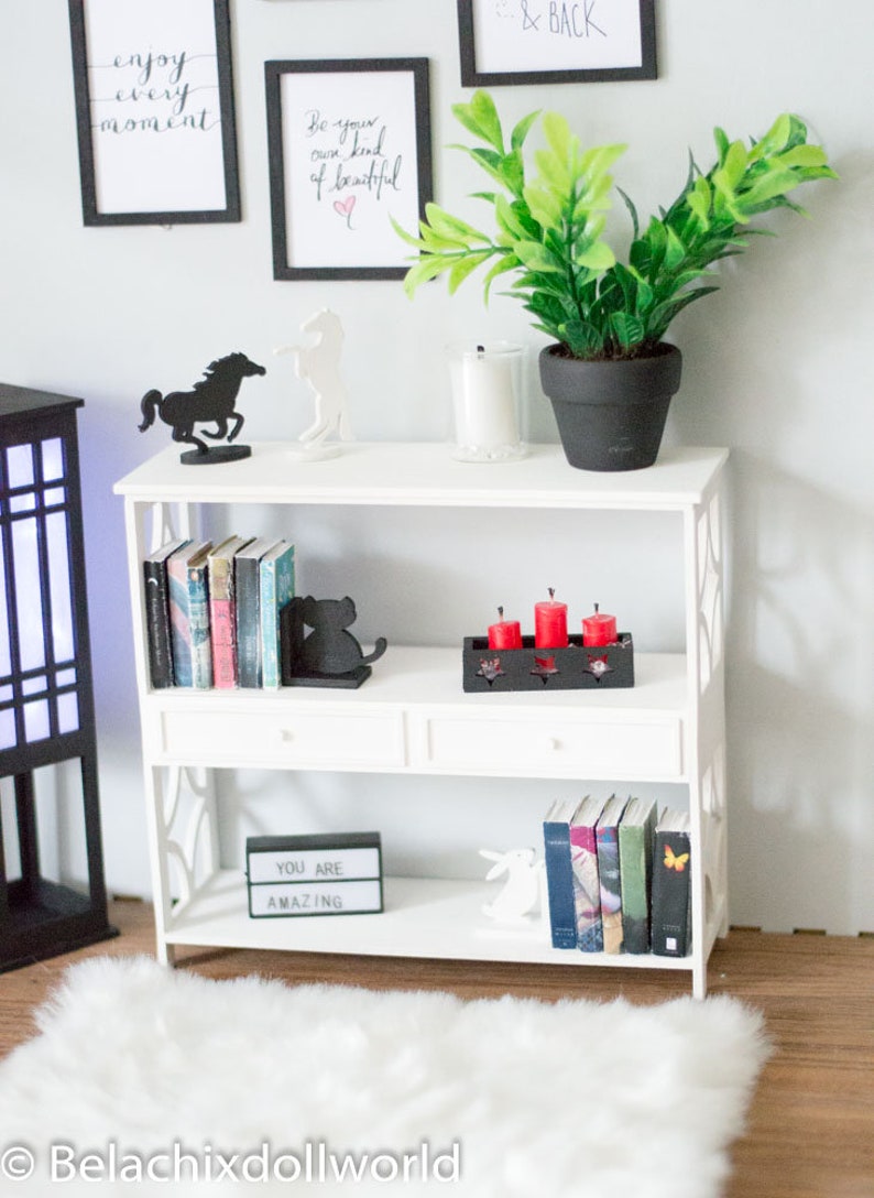 May include: White bookshelf with three shelves, two drawers, and decorative accents. The top shelf has a black horse silhouette, a white horse silhouette, a potted green plant, and a white candle. The middle shelf has a black cat bookend, a set of red candles in a black holder, and a stack of books. The bottom shelf has a black light-up sign that says "You Are Amazing", a white rabbit figurine, and a stack of books.