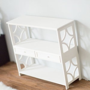 May include: White wooden shelf unit with two shelves and two drawers. The shelf has a decorative cut-out design on the sides.