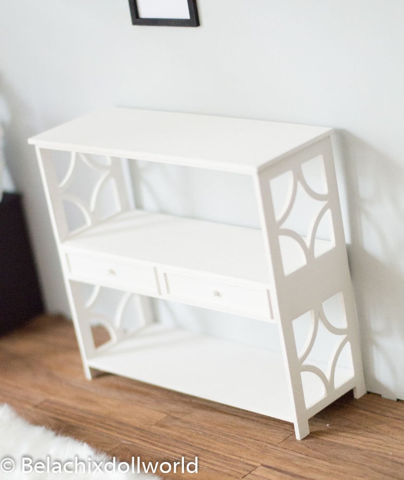 May include: White wooden shelf unit with two shelves and two drawers. The shelf has a decorative cut-out design on the sides.
