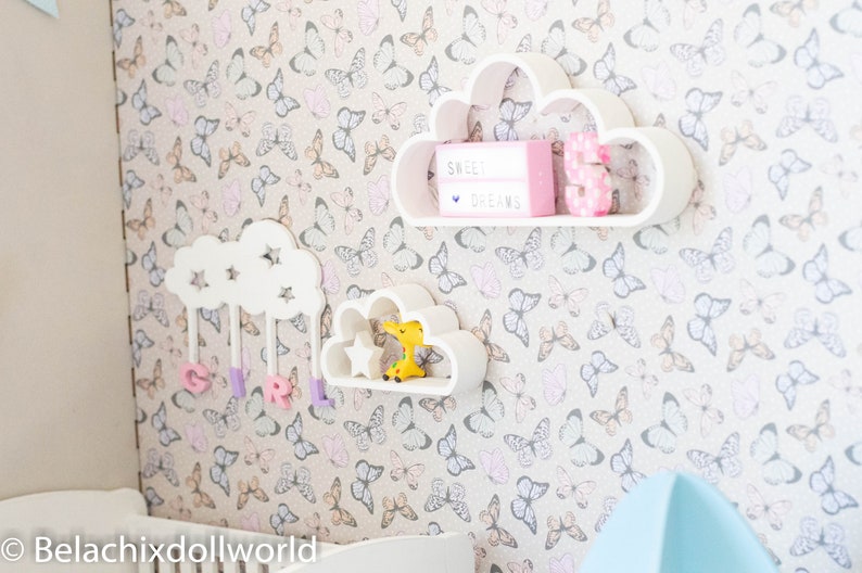 May include: White cloud-shaped shelves with a star design, mounted on a wall with a butterfly pattern wallpaper. The top shelf has a pink light box with the text "Sweet Dreams" and a pink number 3. The middle shelf has a small yellow toy giraffe. The bottom shelf has purple letters spelling "GIRL".