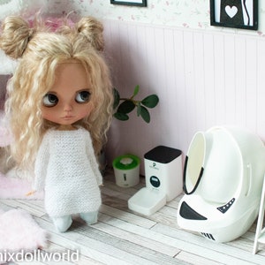 May include: A white doll with blonde curly hair wearing a white sweater and blue pants stands in a dollhouse room. The dollhouse room has a pink cat tree, a white self-cleaning litter box, and a white shelf with plants.