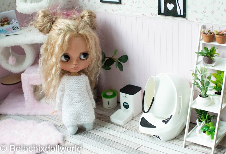 May include: A white doll with blonde curly hair wearing a white sweater and blue pants stands in a dollhouse room. The dollhouse room has a pink cat tree, a white self-cleaning litter box, and a white shelf with plants.