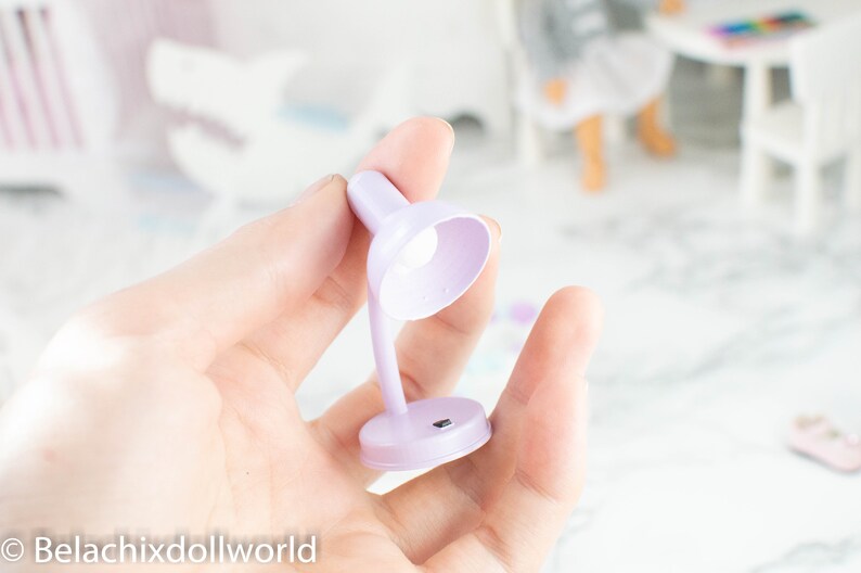 以下が含まれることがあります： 白熱電球が付いた、小さなラベンダー色のデスクランプ。ランプはプラスチック製で、シンプルなデザインです。