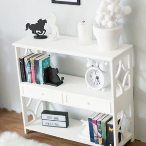 May include: White bookshelf with three shelves and two drawers. The top shelf has a white candle in a glass jar, a white potted plant with white pom poms, and two black silhouette figures of horses. The middle shelf has a row of books, a black silhouette figure of a cat, and a white alarm clock. The bottom shelf has a black sign that says "You are amazing", a row of books, and a white rabbit figurine.