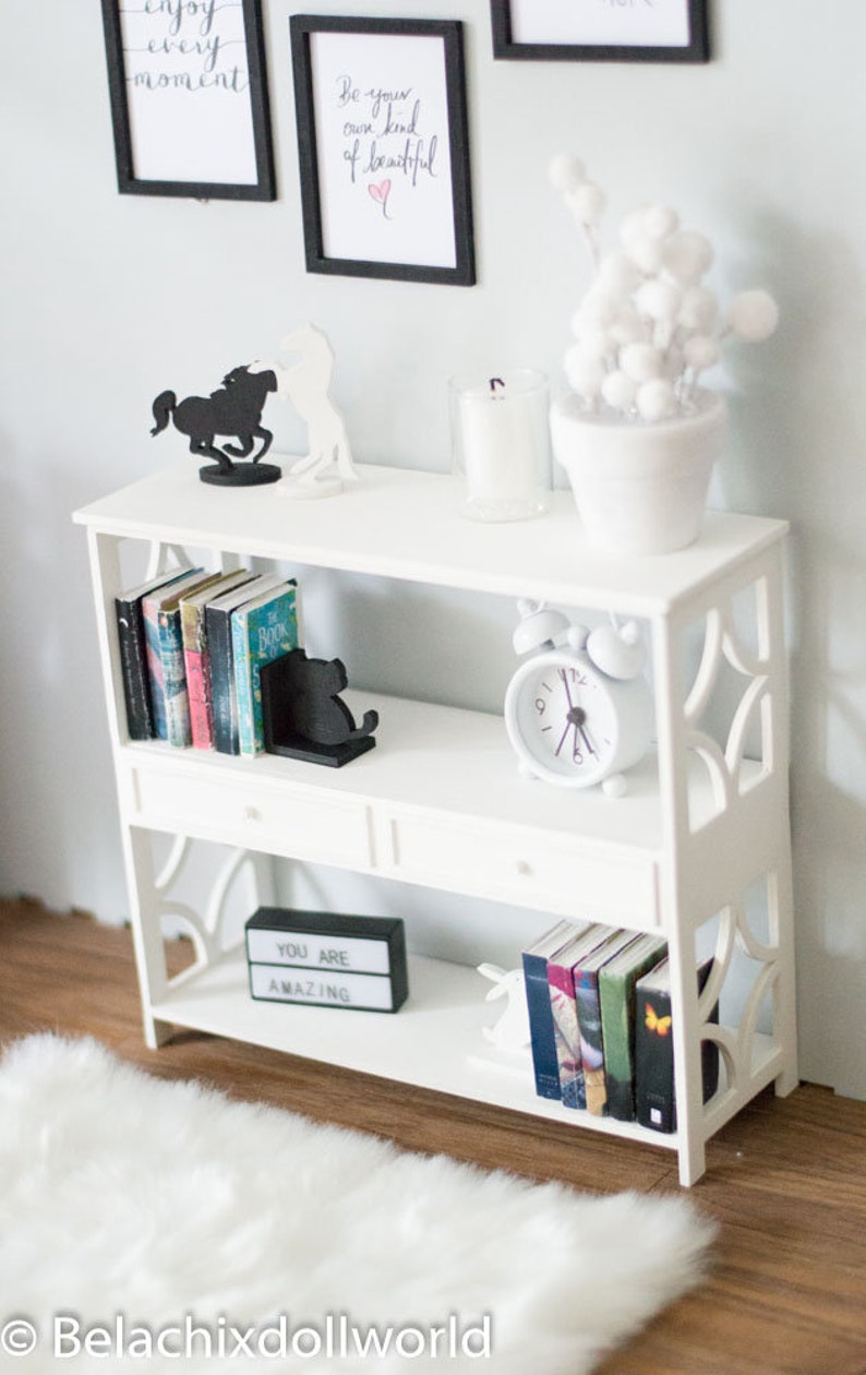 May include: White bookshelf with three shelves and two drawers. The top shelf has a white candle in a glass jar, a white potted plant with white pom poms, and two black silhouette figures of horses. The middle shelf has a row of books, a black silhouette figure of a cat, and a white alarm clock. The bottom shelf has a black sign that says "You are amazing", a row of books, and a white rabbit figurine.