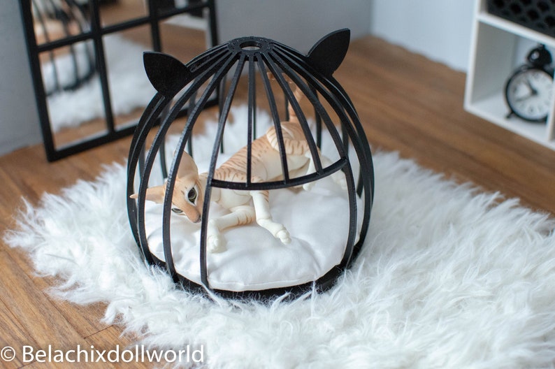 May include: A black cat bed shaped like a cage with a white cushion inside. A tan cat doll is lying inside the bed.