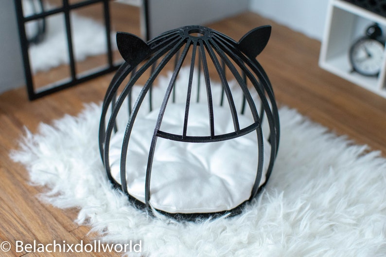 May include: Black cat-shaped bed for small pets, featuring a white cushion and a black wire frame. The bed is on a white fluffy rug.
