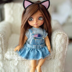 May include: A doll with brown hair and blue eyes wearing a denim dress and a cat ear headband. The doll is sitting on a white knitted chair. The doll's outfit includes a light blue top and a denim overall dress.