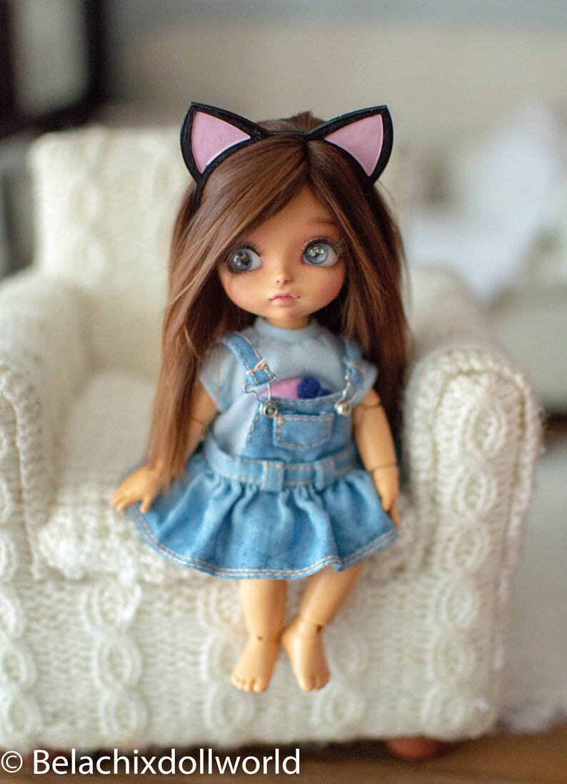 May include: A doll with brown hair and blue eyes wearing a denim dress and a cat ear headband. The doll is sitting on a white knitted chair. The doll's outfit includes a light blue top and a denim overall dress.