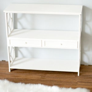 May include: White wooden shelf unit with two drawers and open shelves. The shelf unit is on a wood floor with a white fluffy rug in front of it.