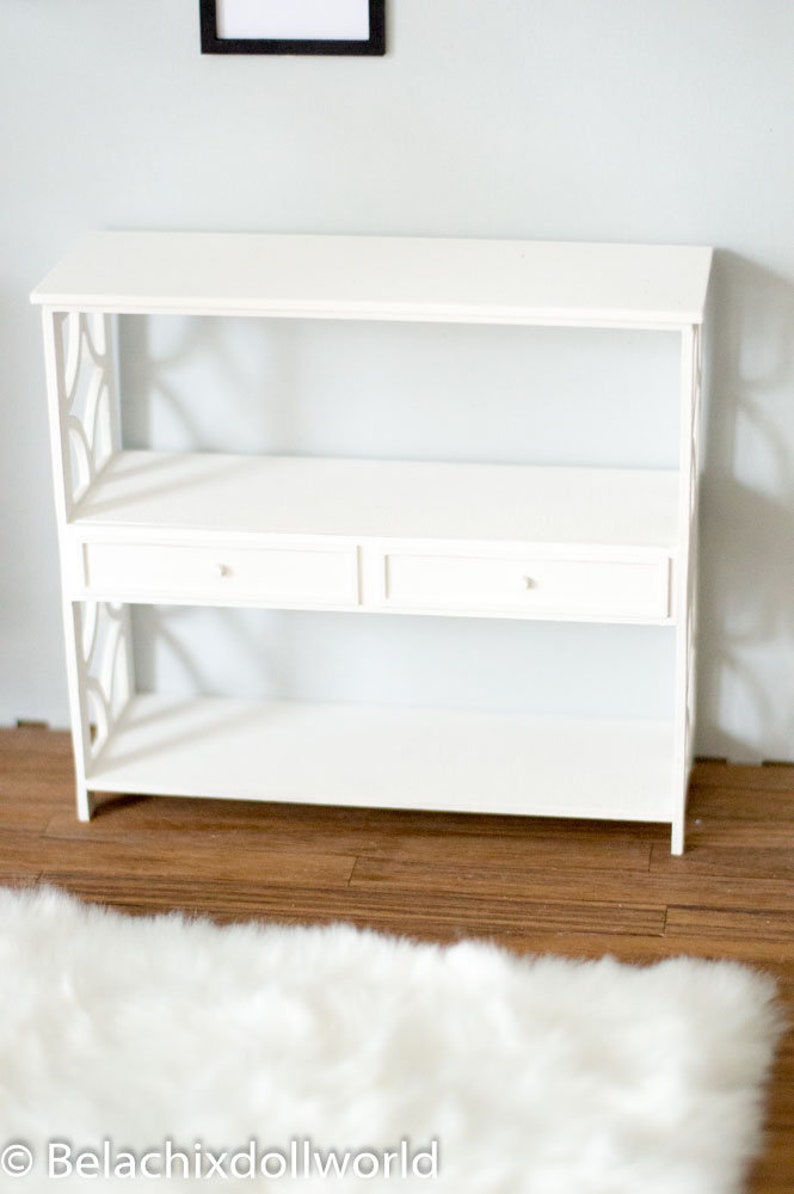 May include: White wooden shelf unit with two drawers and open shelves. The shelf unit is on a wood floor with a white fluffy rug in front of it.