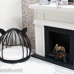 May include: Black metal cat bed with a white cushion inside, next to a white fireplace with a black brick surround and a lit fire inside.