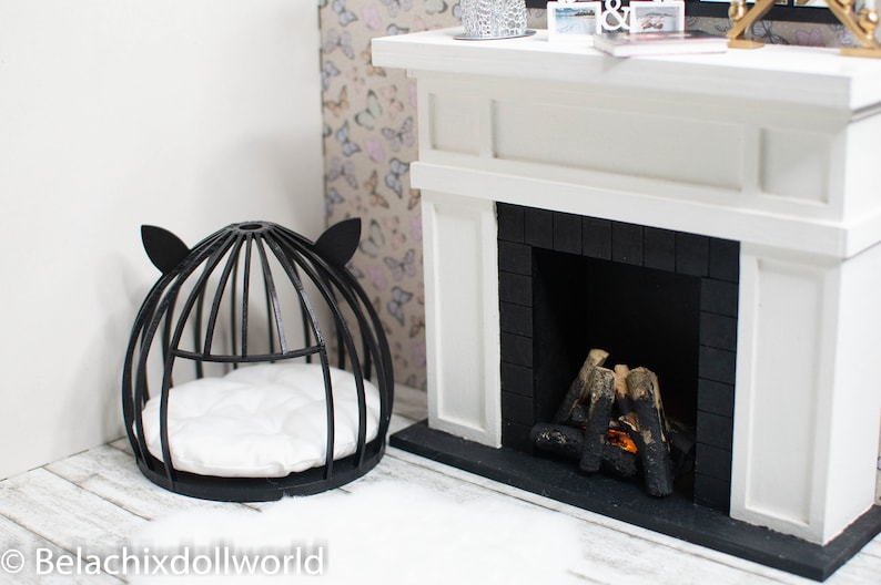 May include: Black metal cat bed with a white cushion inside, next to a white fireplace with a black brick surround and a lit fire inside.