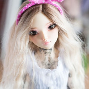 May include: A doll with long, wavy, blonde hair wearing a pink polka dot headband and a white dress with a lace trim. The doll has a delicate face with blue eyes and a light pink blush.