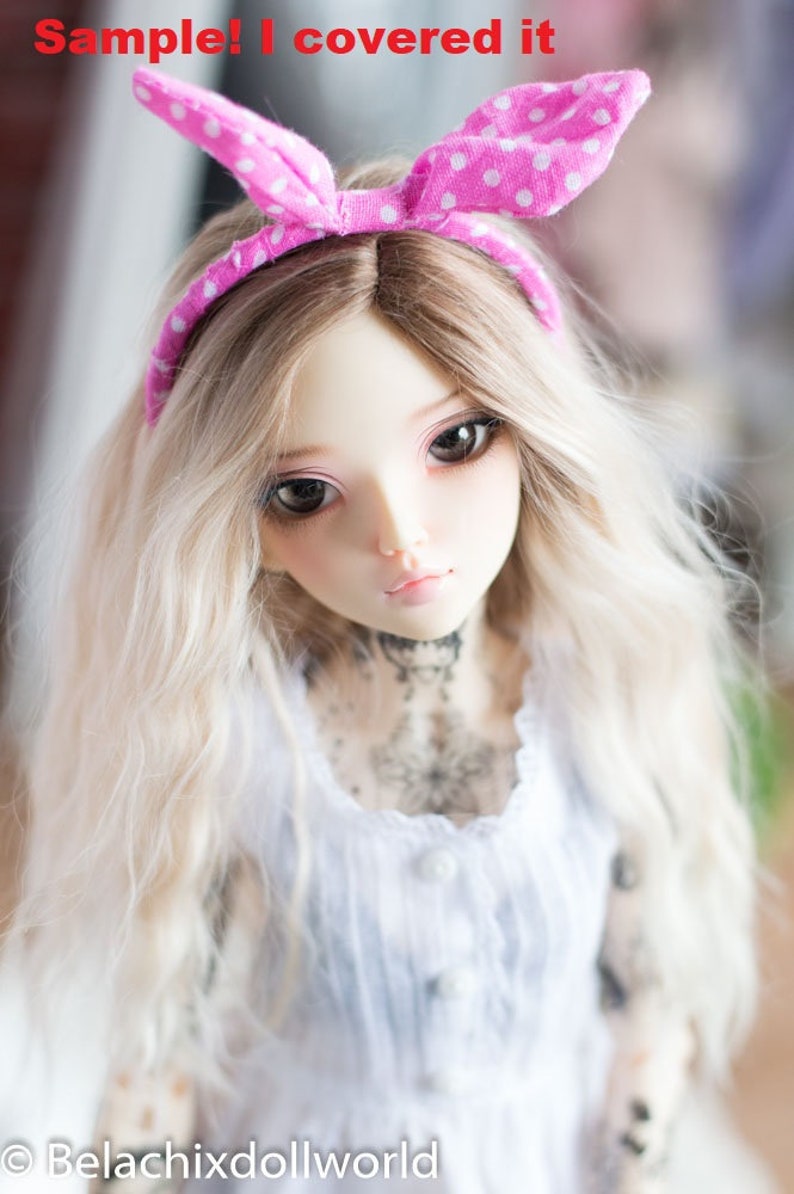 May include: A doll with long, wavy, blonde hair wearing a pink polka dot headband and a white dress with a lace trim. The doll has a delicate face with blue eyes and a light pink blush.