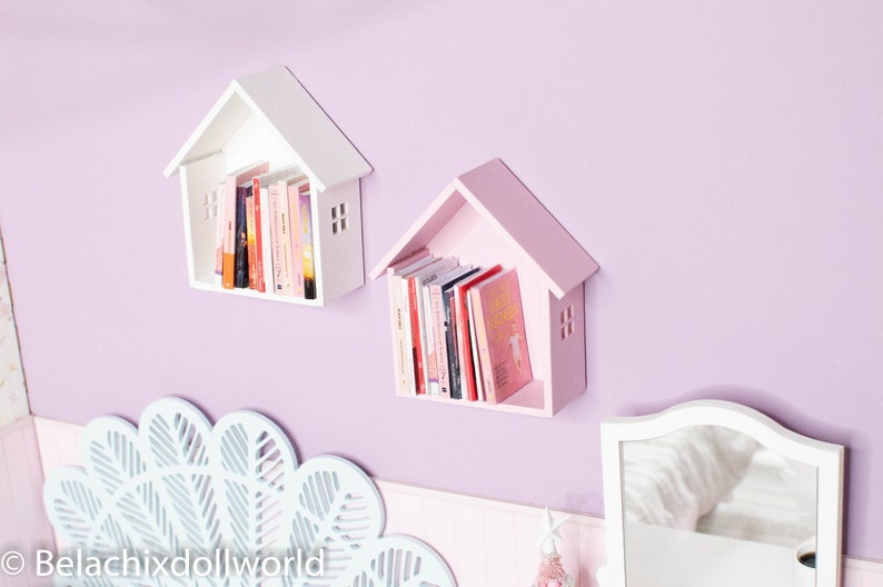 May include: Two small house-shaped bookshelves, one white and one pink, mounted on a light purple wall. Both shelves are filled with books.