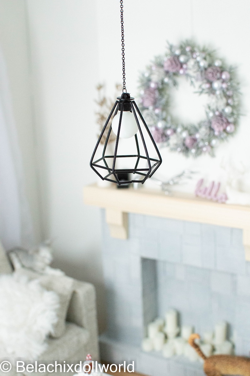 May include: A black metal cage pendant light with a white light bulb hanging from a chain. The light is hanging in front of a white brick fireplace with a wreath on the wall behind it.