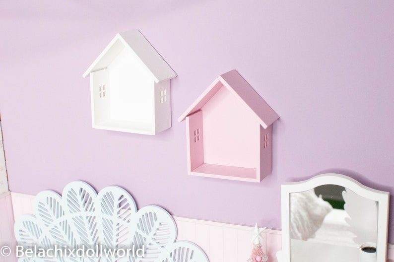 May include: Two small house-shaped shelves, one white and one pink, mounted on a lavender wall. The shelves are empty and have a simple design.
