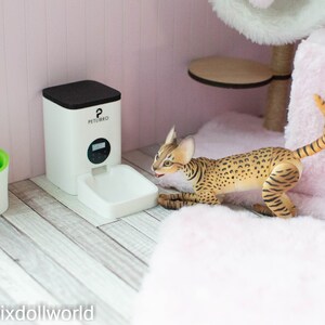 May include: A white automatic pet feeder with a black top and a digital display. The feeder is on a light wood floor. A small green water fountain with a white daisy on top is to the left of the feeder. A brown and tan cat is lying on a pink fluffy bed.