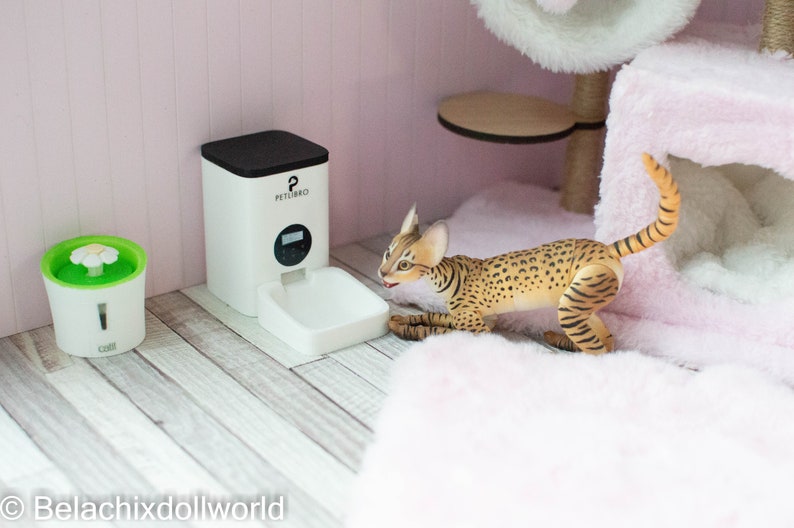 May include: A white automatic pet feeder with a black top and a digital display. The feeder is on a light wood floor. A small green water fountain with a white daisy on top is to the left of the feeder. A brown and tan cat is lying on a pink fluffy bed.