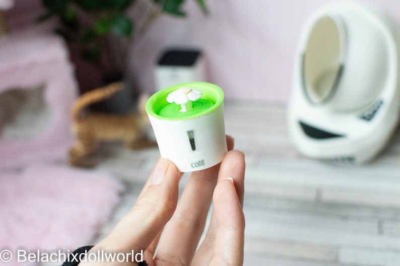 May include: A small, white and green cat water fountain with a white flower on top. The fountain is labeled "Catit".