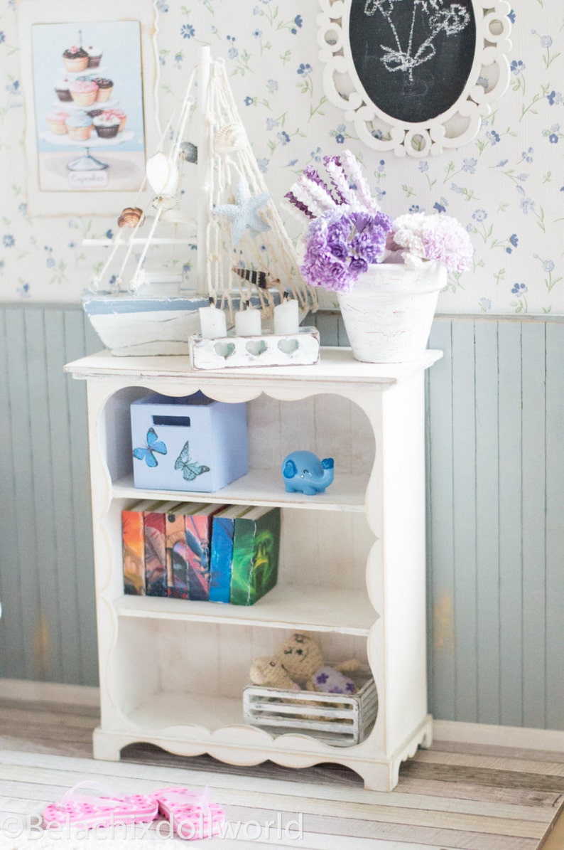 Pu&ograve; includere: Libreria bianca a tre ripiani, con una scatola blu con adesivi a forma di farfalla, una statuetta di elefante blu, un set di libri colorati, una cassa bianca con un orsacchiotto e un fiore viola, e una base bianca con un bordo a festoni.