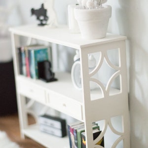 May include: White bookshelf with three shelves and a drawer. The bookshelf has a decorative cut-out design on the front. The top shelf has a white planter with a white fluffy plant. The middle shelf has books and a black decorative object. The bottom shelf has books.