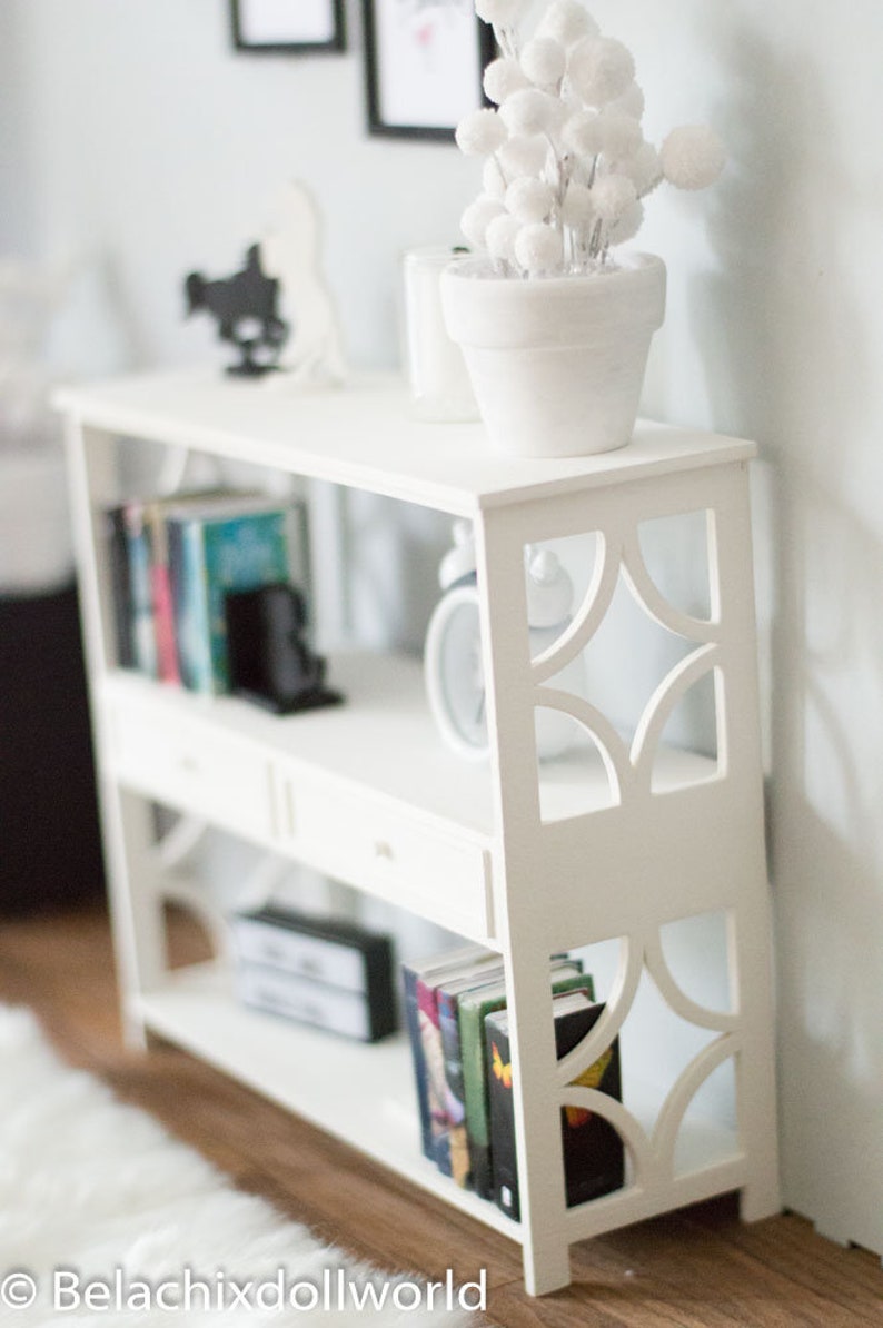 May include: White bookshelf with three shelves and a drawer. The bookshelf has a decorative cut-out design on the front. The top shelf has a white planter with a white fluffy plant. The middle shelf has books and a black decorative object. The bottom shelf has books.