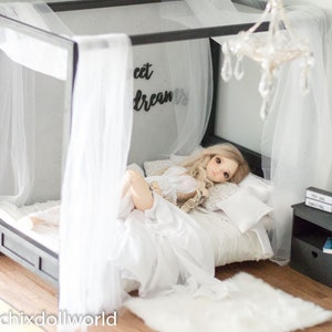 Puede incluir: Una mu&ntilde;eca blanca con cabello rubio yace en una cama con dosel blanco con un edred&oacute;n y almohadas blancas. La cama tiene un marco de metal negro y las palabras "Sweet Dreams" est&aacute;n en la pared detr&aacute;s de la cama.