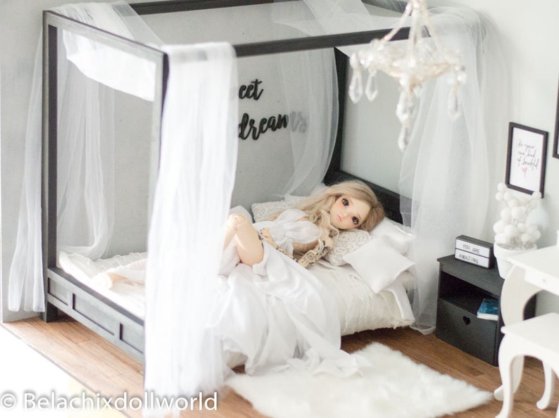 Puede incluir: Una mu&ntilde;eca blanca con cabello rubio yace en una cama con dosel blanco con un edred&oacute;n y almohadas blancas. La cama tiene un marco de metal negro y las palabras "Sweet Dreams" est&aacute;n en la pared detr&aacute;s de la cama.