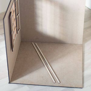 May include: A dollhouse room with beige walls and a wooden window. Two wooden strips are lying on the floor.