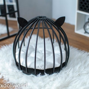 May include: Black cat-shaped bed for small pets, featuring a round, wire frame with a white cushion inside. The bed is on a white fluffy rug.