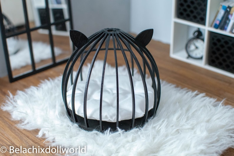 May include: Black cat-shaped bed for small pets, featuring a round, wire frame with a white cushion inside. The bed is on a white fluffy rug.