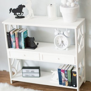 May include: White bookshelf with three shelves and two drawers. The top shelf has two black metal horse silhouettes, a white candle in a glass jar, and a white planter with white pom poms. The middle shelf has a set of books with black bookends shaped like a cat, and a white alarm clock. The bottom shelf has a black sign that says "You are amazing", a set of books, and a white rabbit figurine.