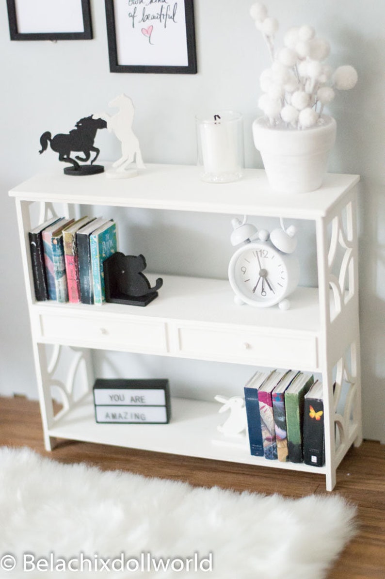May include: White bookshelf with three shelves and two drawers. The top shelf has two black metal horse silhouettes, a white candle in a glass jar, and a white planter with white pom poms. The middle shelf has a set of books with black bookends shaped like a cat, and a white alarm clock. The bottom shelf has a black sign that says "You are amazing", a set of books, and a white rabbit figurine.