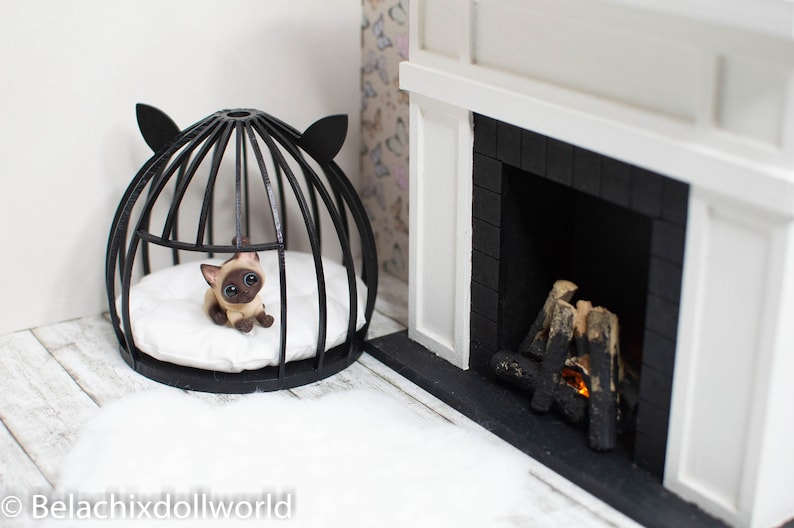May include: A black wire cat bed with a white cushion and a small Siamese cat figurine inside. The bed is in front of a white fireplace with a black brick surround and a small fire inside.