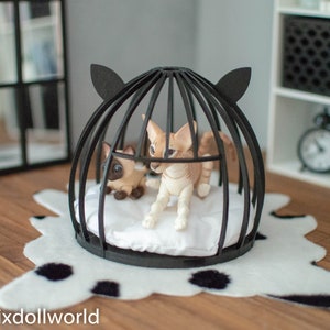 May include: Two small, white and brown, cat figurines inside a black wire cage with a white cushion. The cage is on a white and black spotted rug.