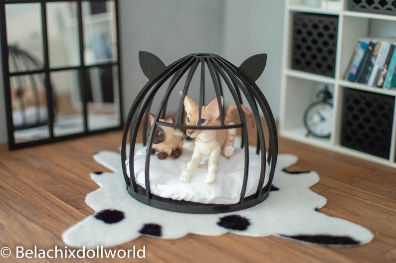 May include: Two small, white and brown, cat figurines inside a black wire cage with a white cushion. The cage is on a white and black spotted rug.