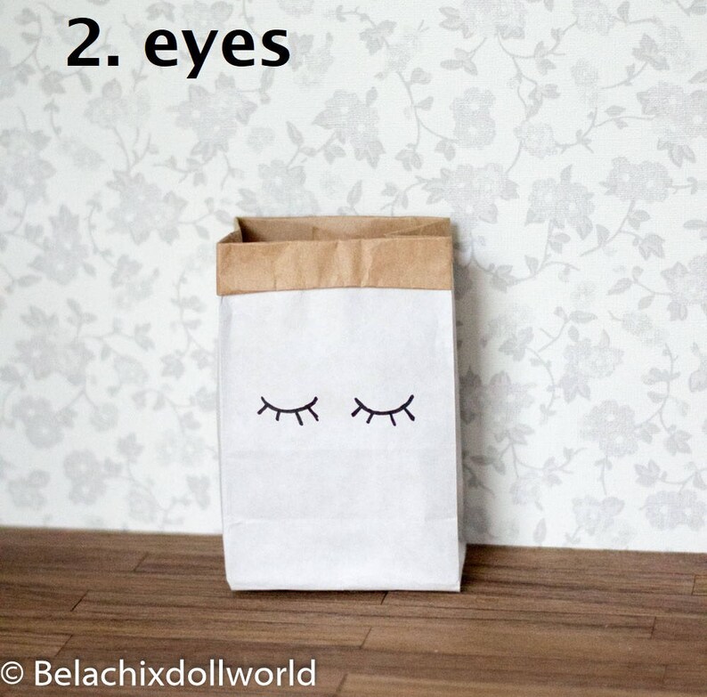 May include: A white paper bag with brown paper trim and two black eyes drawn on the front. The text "2. eyes" is written above the bag.