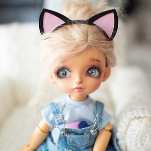 May include: A doll with large blue eyes and blonde hair, wearing a denim overall dress and a light blue shirt. The doll has a black and pink cat ear headband. The doll's face is painted with detailed features. The doll is a collectible item.