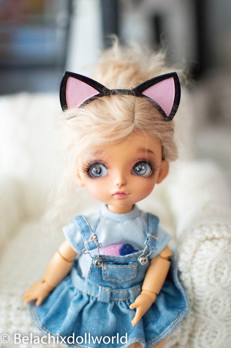 May include: A doll with large blue eyes and blonde hair, wearing a denim overall dress and a light blue shirt. The doll has a black and pink cat ear headband. The doll's face is painted with detailed features. The doll is a collectible item.