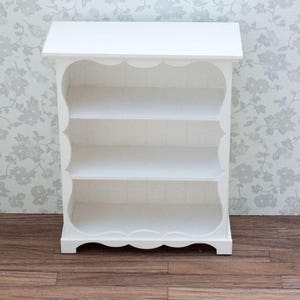 Pu&ograve; includere: Libreria in legno bianco con tre ripiani e un bordo superiore e inferiore a forma di conchiglia. La libreria &egrave; appoggiata su un pavimento in legno.