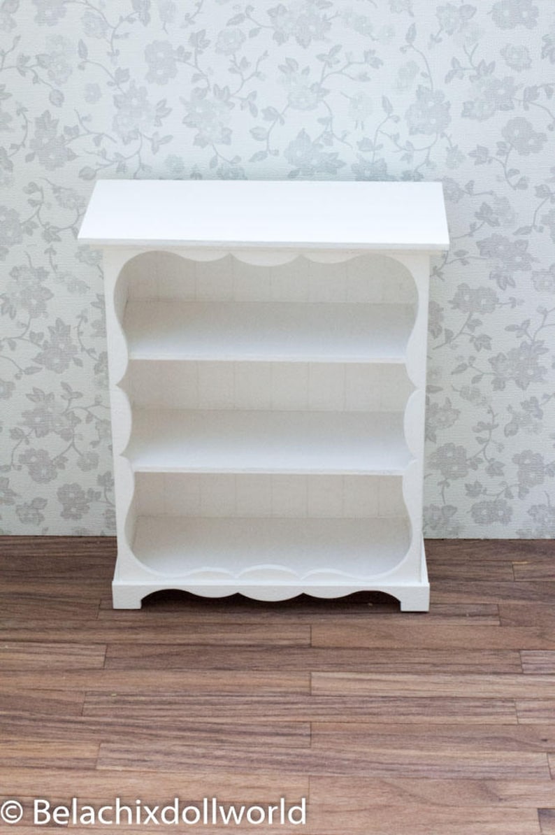 Pu&ograve; includere: Libreria in legno bianco con tre ripiani e un bordo superiore e inferiore a forma di conchiglia. La libreria &egrave; appoggiata su un pavimento in legno.